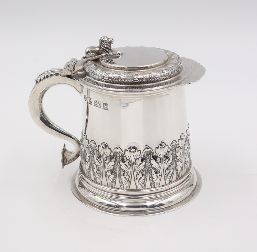 A sterling silver lidded tankard featuring chased acanthus leaf decoration, a scroll handle, visible hallmarks, and a lion thumbpiece.