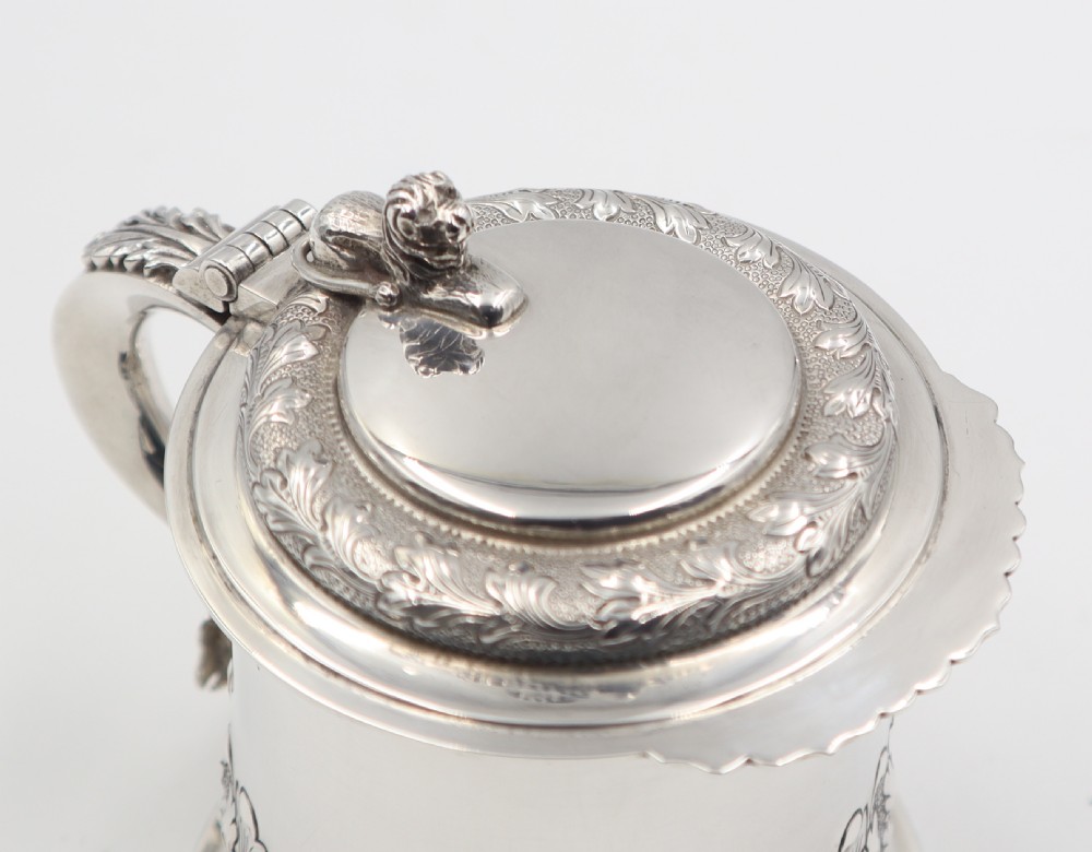 A sterling silver lidded tankard featuring chased acanthus leaf decoration, a scroll handle, visible hallmarks, and a lion thumbpiece. View V