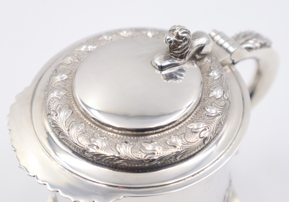 A sterling silver lidded tankard featuring chased acanthus leaf decoration, a scroll handle, visible hallmarks, and a lion thumbpiece. View VI
