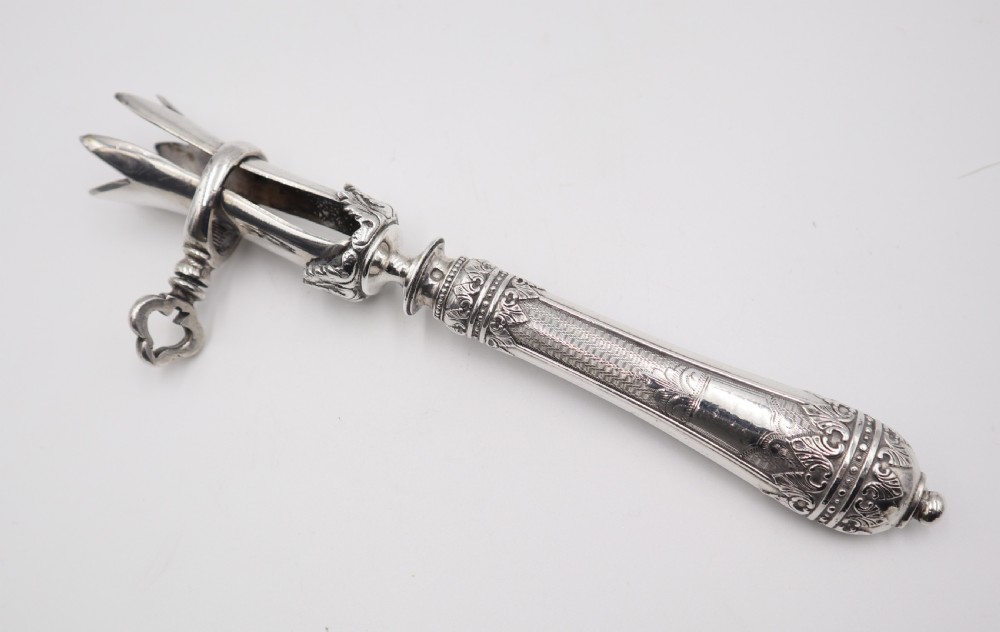 An antique silver ham bone holder featuring a decorated handle with chased leaf motifs, a threaded key screw, and adjustable pronged clamps. View IV