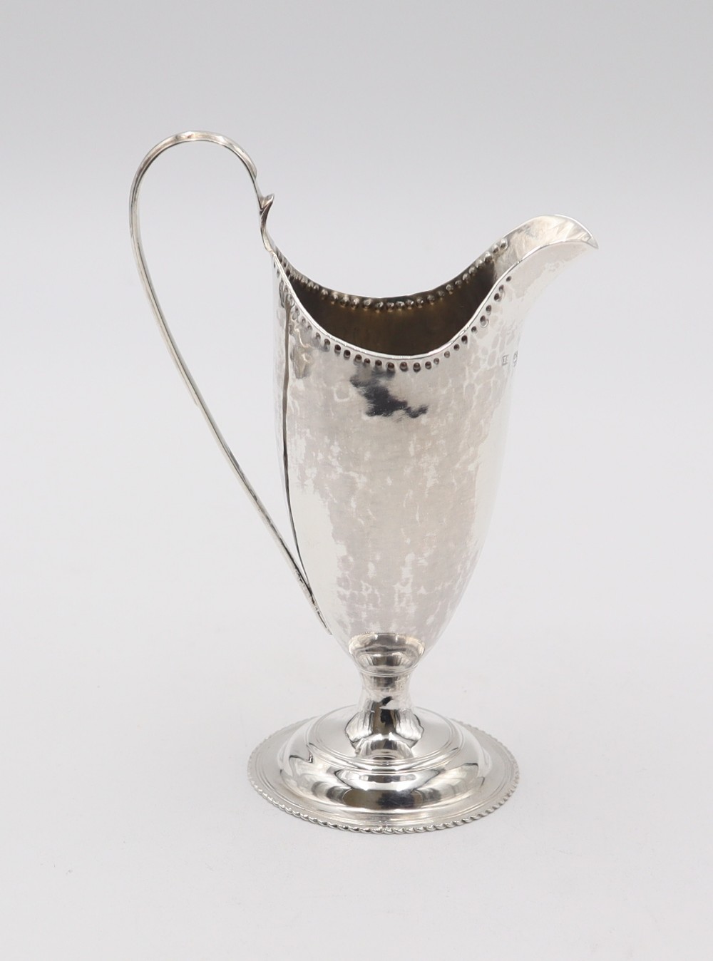 A hand-hammered sterling silver cream jug featuring a high loop handle, beaded rim, and circular pedestal base.