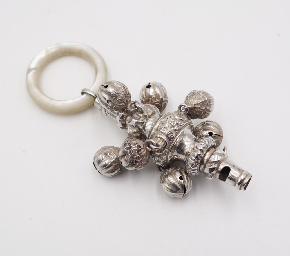An antique silver baby rattle featuring embossed floral scrollwork, attached jingle bells, an integrated whistle end, and a smooth circular mother of pearl ring handle.