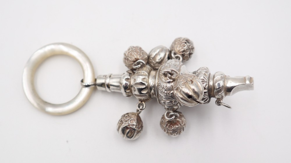 An antique silver baby rattle featuring embossed floral scrollwork, attached jingle bells, an integrated whistle end, and a smooth circular mother of pearl ring handle. View II
