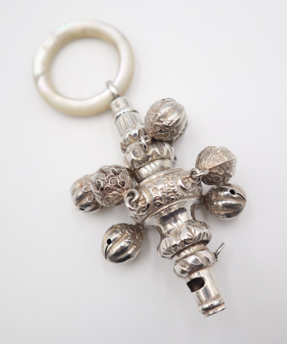 An antique silver baby rattle featuring embossed floral scrollwork, attached jingle bells, an integrated whistle end, and a smooth circular mother of pearl ring handle. View IV