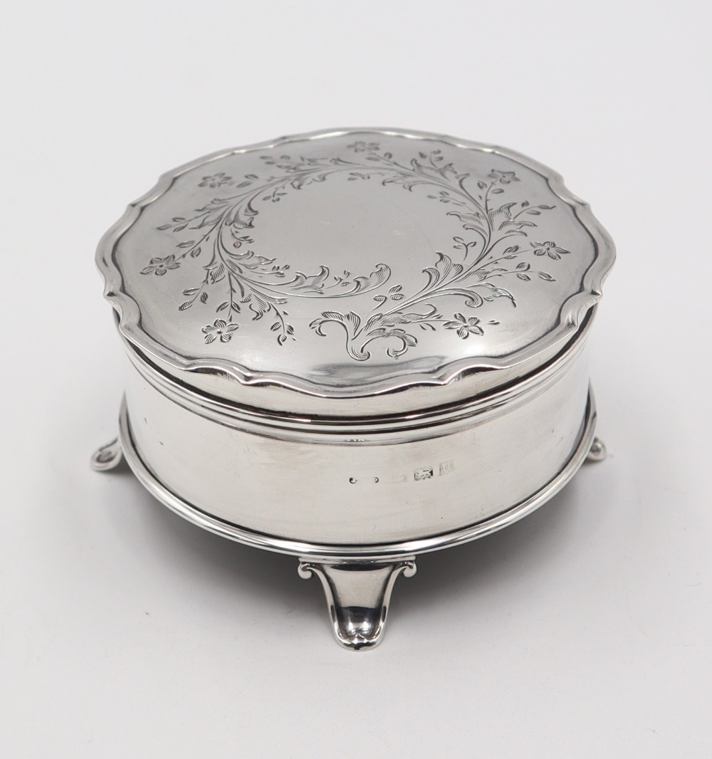 An antique round silver trinket box featuring an engraved floral leaf top lid, a scalloped border, and four cabriole feet.