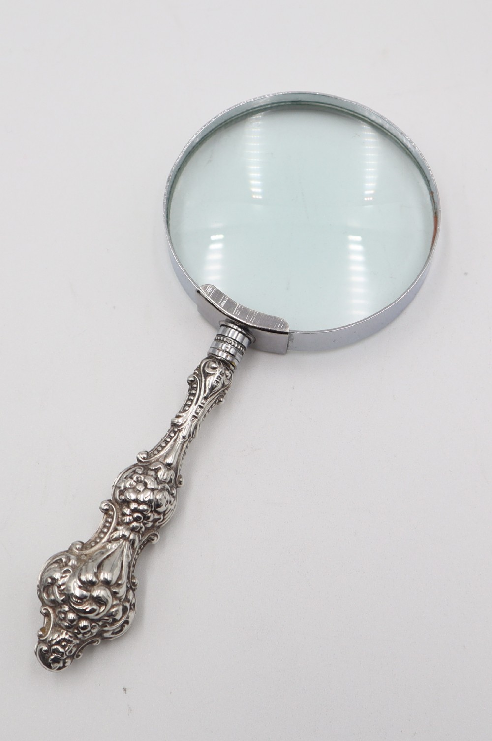 An antique hand-held magnifying glass with a circular lens and a decorative repousse silver handle featuring scroll patterns.