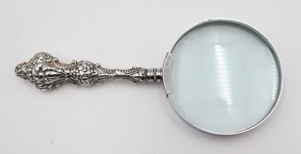 An antique hand-held magnifying glass with a circular lens and a decorative repousse silver handle featuring scroll patterns. View II