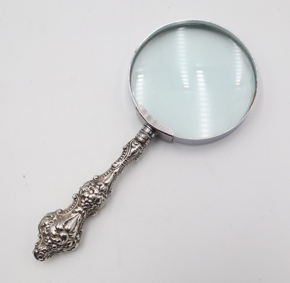 An antique hand-held magnifying glass with a circular lens and a decorative repousse silver handle featuring scroll patterns. View III