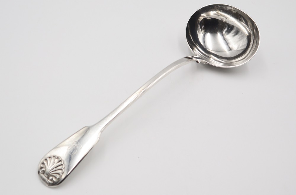 An antique silver soup ladle featuring a deep circular bowl, a slender arched stem, and a shell detail on the handle terminal.