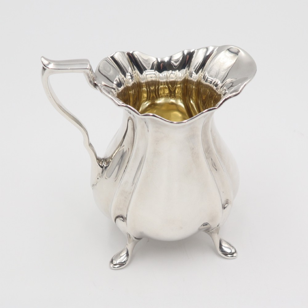 An antique silver cream jug featuring a lobed body, wavy rim, single scroll handle, three pad feet, and a gilded interior.