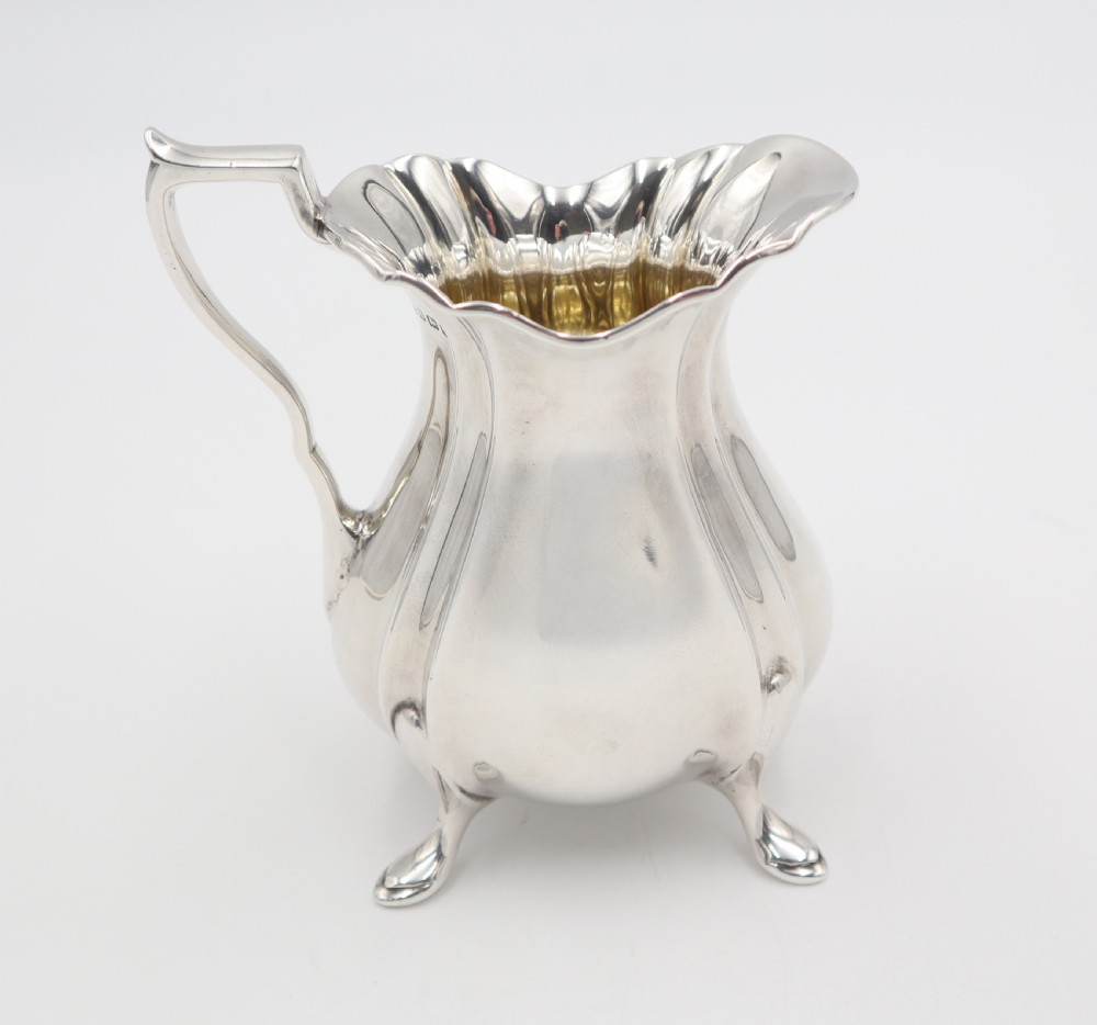 An antique silver cream jug featuring a lobed body, wavy rim, single scroll handle, three pad feet, and a gilded interior. View II