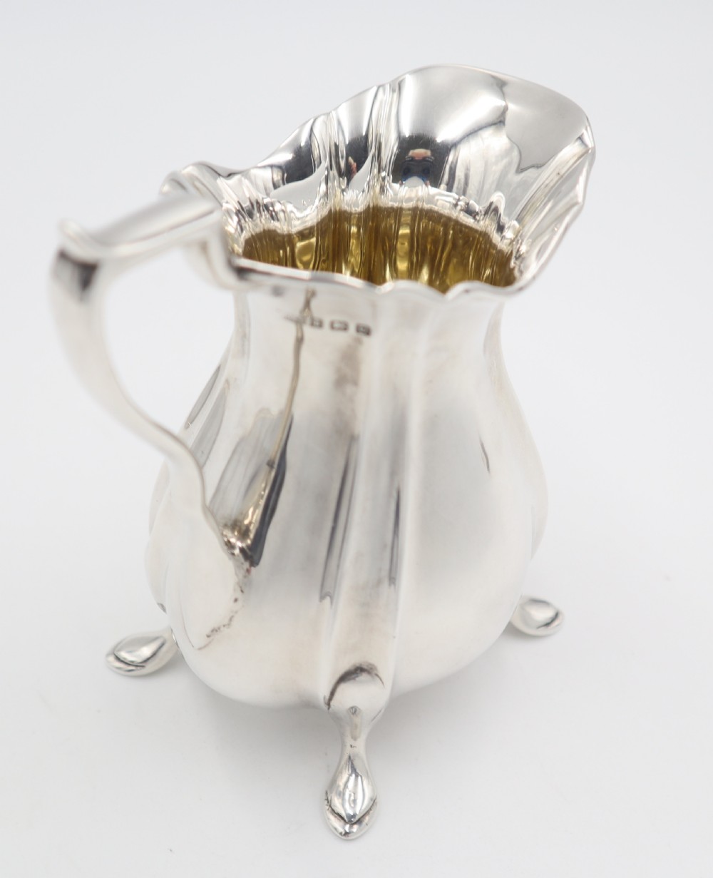 An antique silver cream jug featuring a lobed body, wavy rim, single scroll handle, three pad feet, and a gilded interior. View IV
