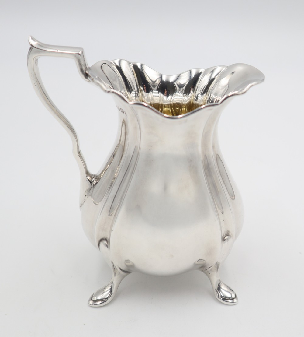 An antique silver cream jug featuring a lobed body, wavy rim, single scroll handle, three pad feet, and a gilded interior. View V