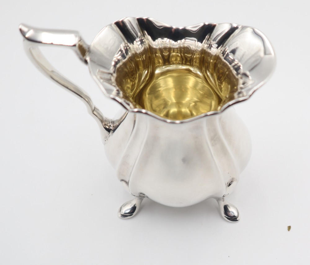 An antique silver cream jug featuring a lobed body, wavy rim, single scroll handle, three pad feet, and a gilded interior. View VI
