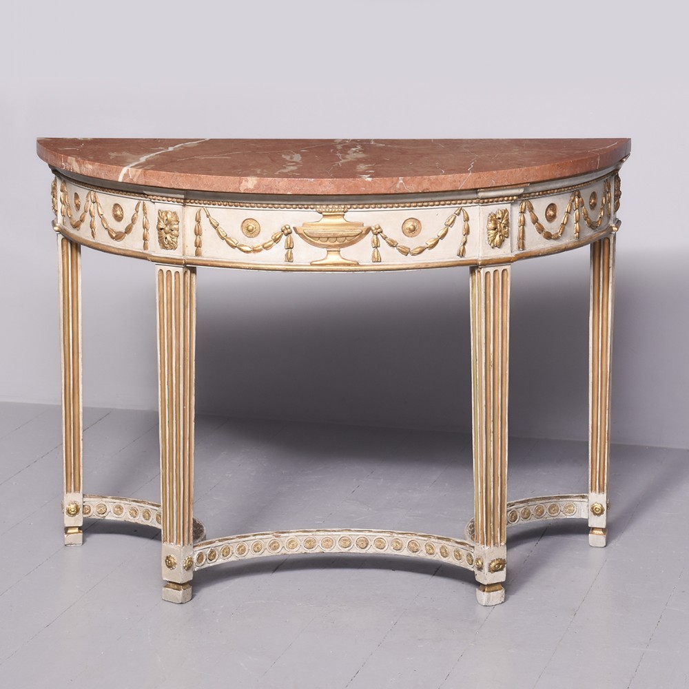 An Adam style semi-circular demi-lune console table featuring a reddish-pink marble top, cream painted frame with gilded urn and swag carvings, fluted legs, and a curved stretcher base.