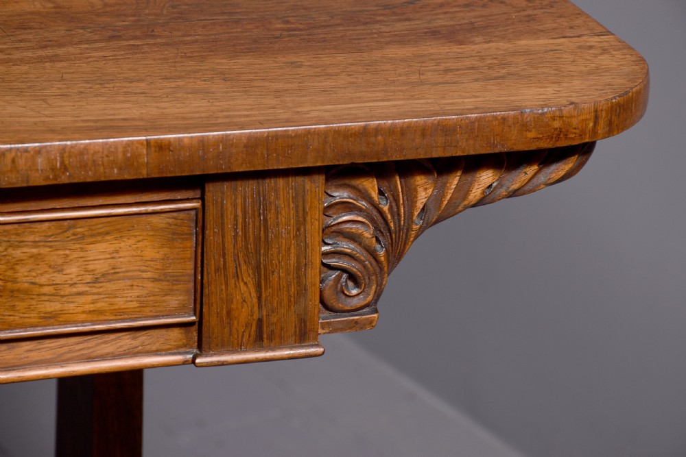 A William IV rosewood library table featuring a rectangular top, two frieze drawers, carved scroll brackets, end pedestal supports, and a turned central stretcher. View IV