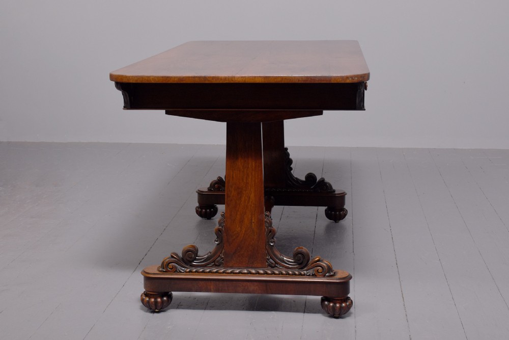 A William IV rosewood library table featuring a rectangular top, two frieze drawers, carved scroll brackets, end pedestal supports, and a turned central stretcher. View VII