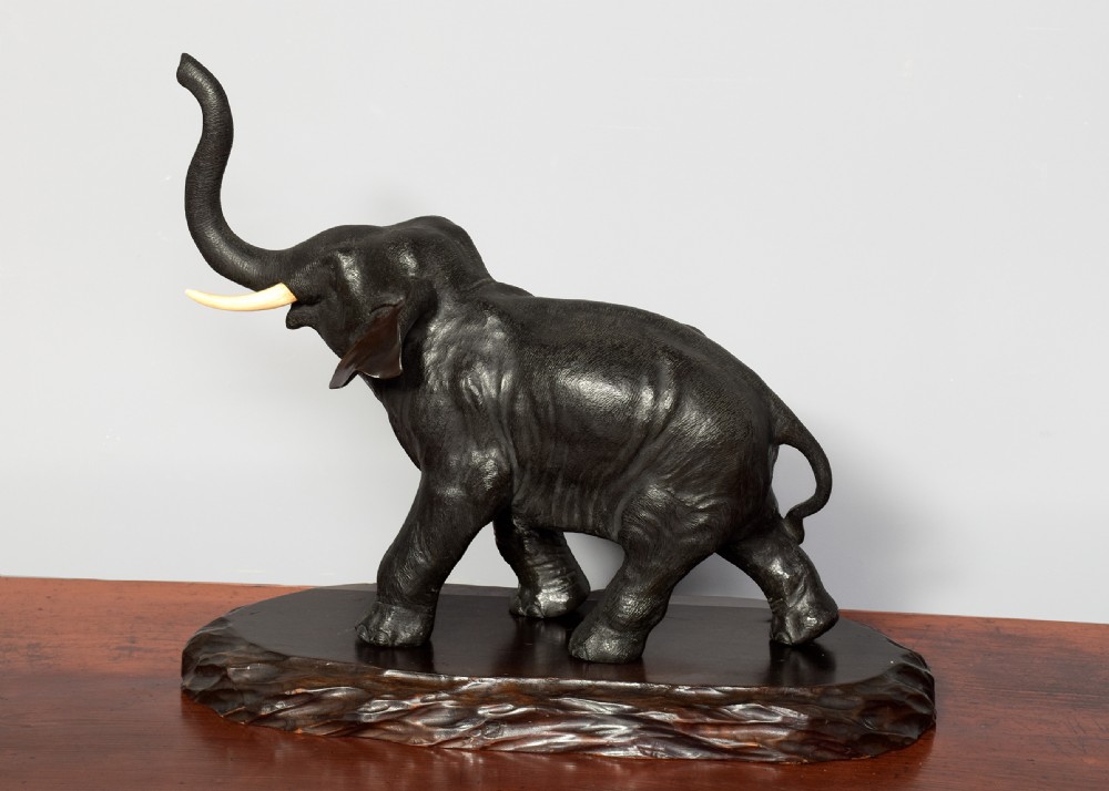 A Japanese dark patinated bronze elephant sculpture with a raised trunk and ivory tusks standing on a naturalistic base. View II