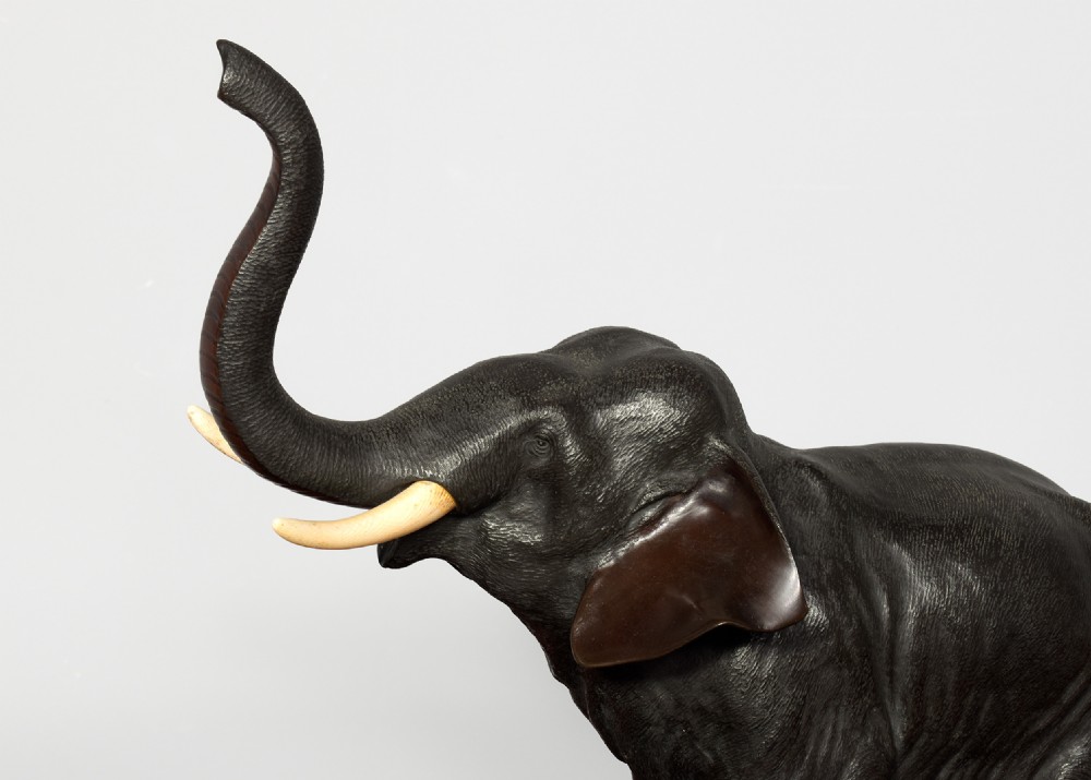 A Japanese dark patinated bronze elephant sculpture with a raised trunk and ivory tusks standing on a naturalistic base. View III