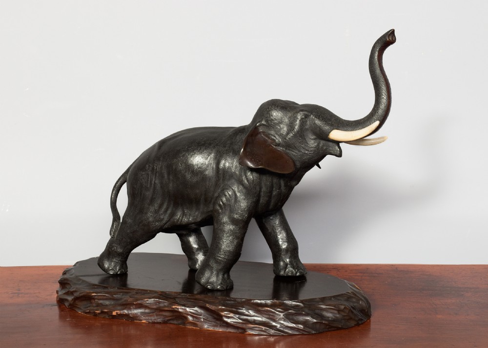 A Japanese dark patinated bronze elephant sculpture with a raised trunk and ivory tusks standing on a naturalistic base. View V