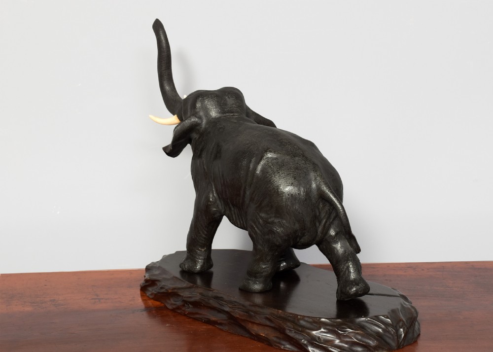 A Japanese dark patinated bronze elephant sculpture with a raised trunk and ivory tusks standing on a naturalistic base. View VI