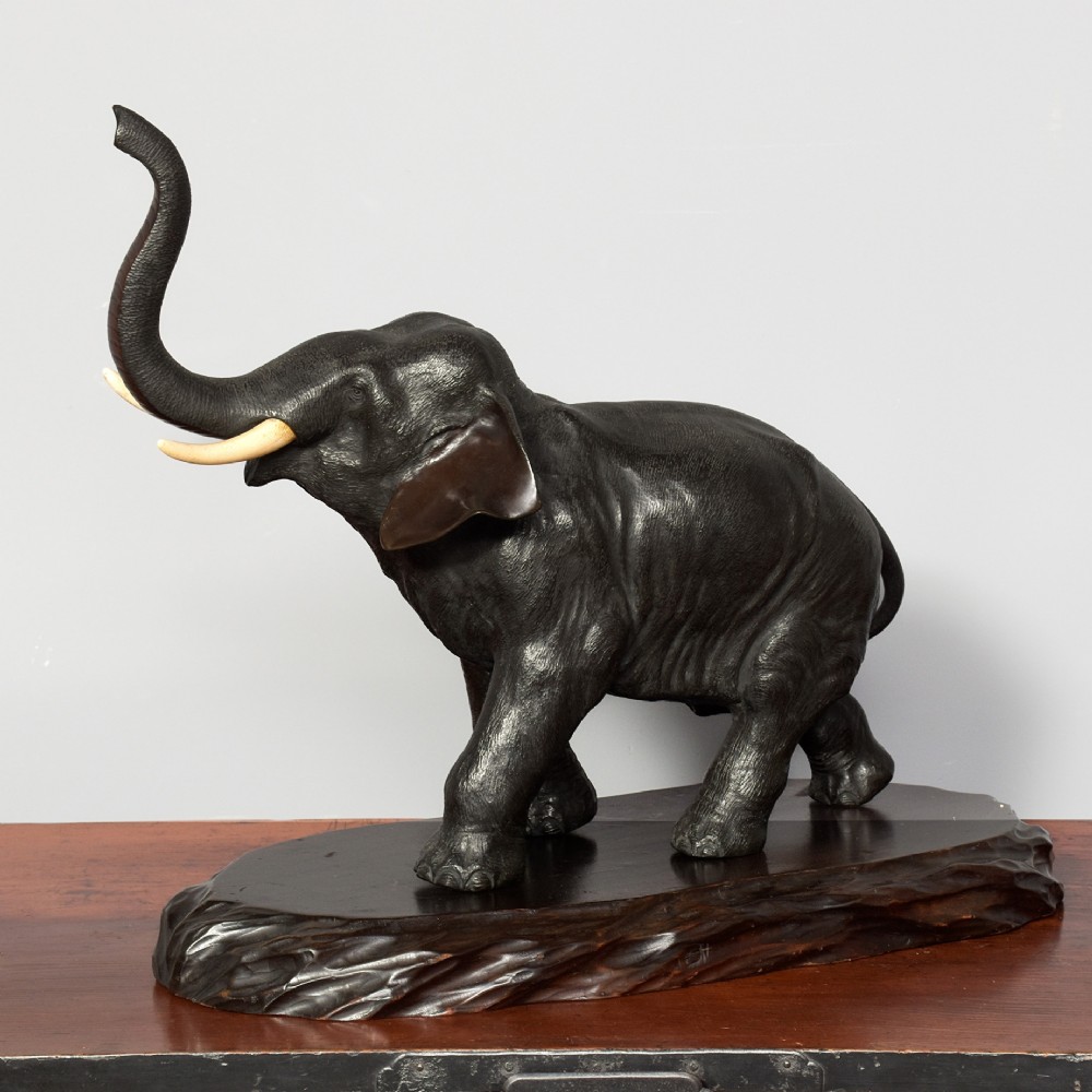 A Japanese dark patinated bronze elephant sculpture with a raised trunk and ivory tusks standing on a naturalistic base.
