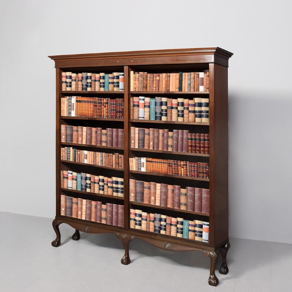 A wide dark mahogany open bookcase featuring twin double bays with multiple shelves, decorative inlay on the frieze, carved shell motifs on the lower apron, and cabriole legs ending in ball and claw feet.