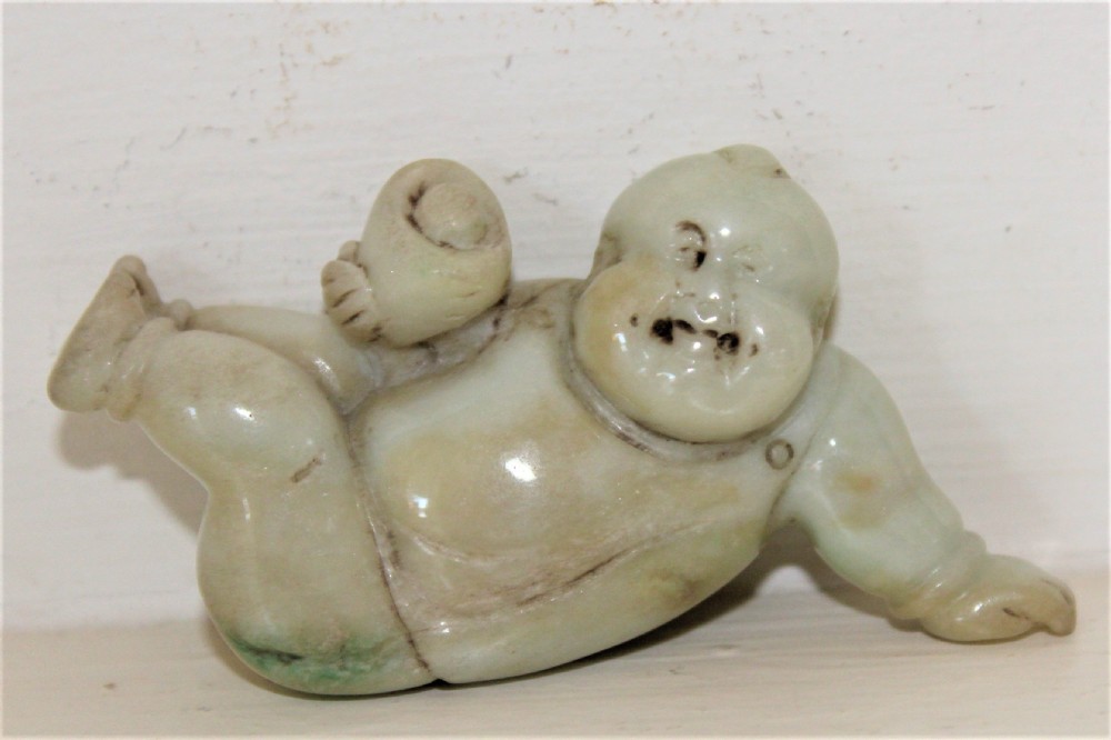 A small carved celadon green jade figure depicting a reclining boy holding a bird nest containing an egg.