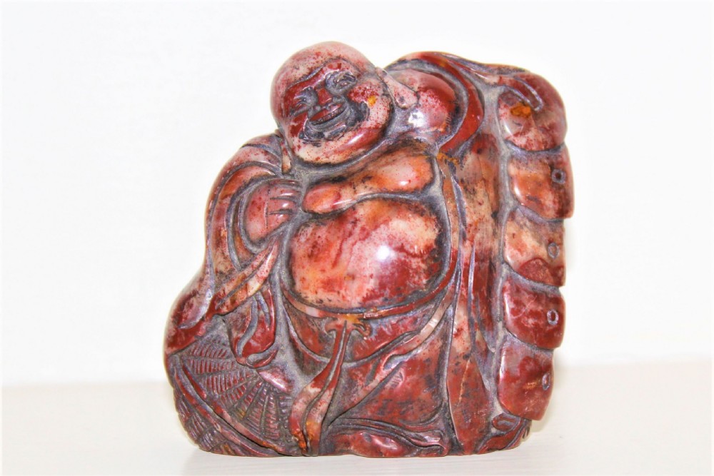 A carved reddish-brown soapstone desk seal depicting a seated, smiling Buddha figure with draped robes. View VI