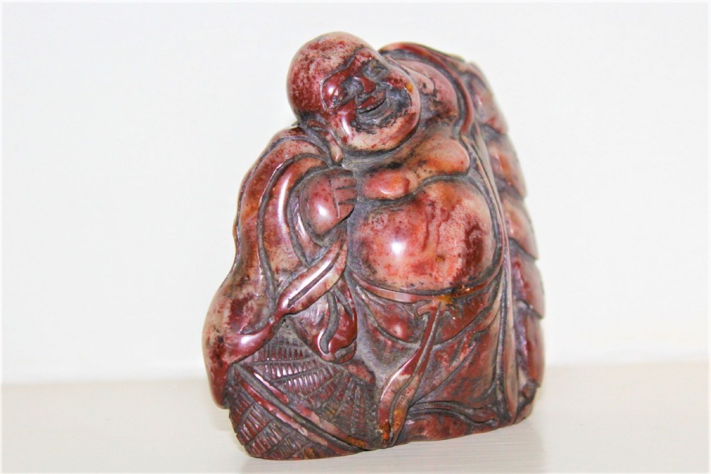 A carved reddish-brown soapstone desk seal depicting a seated, smiling Buddha figure with draped robes. View IV