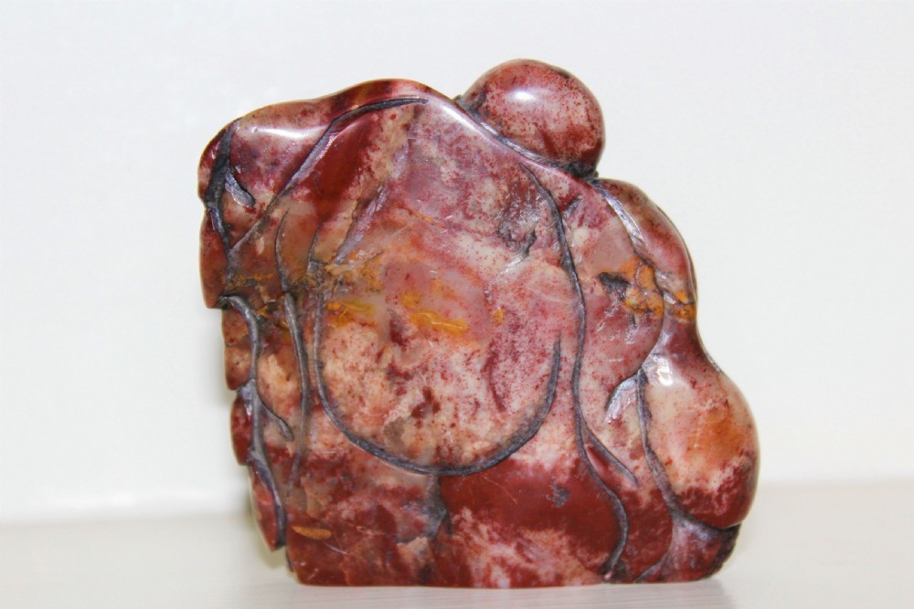 A carved reddish-brown soapstone desk seal depicting a seated, smiling Buddha figure with draped robes. View V