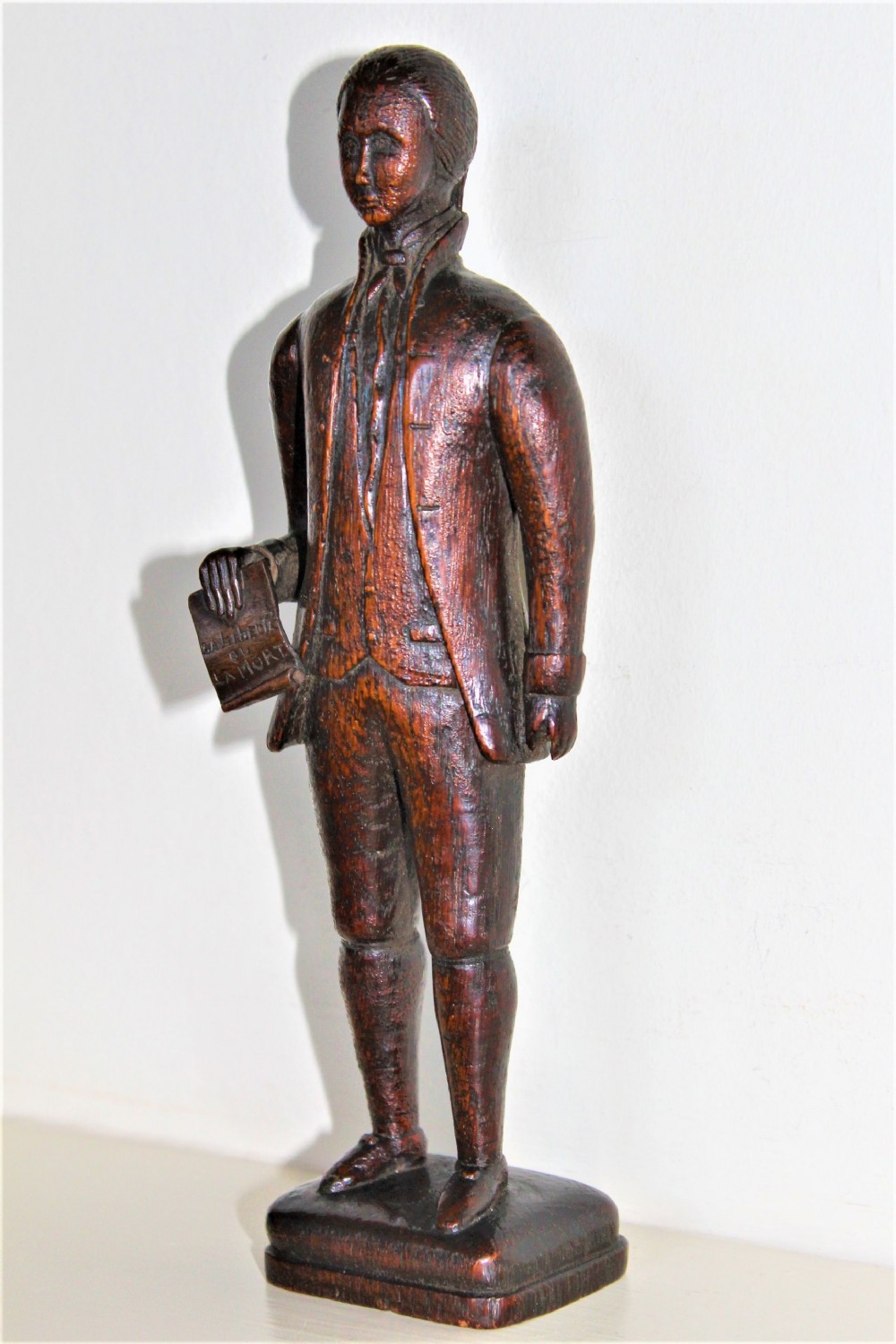 A hand-carved dark wood statuette depicting a male figure in 18th-century clothing holding a scroll on an integrated plinth.