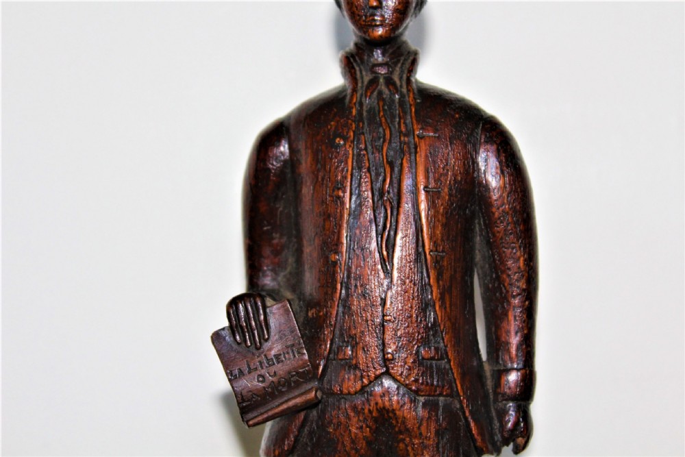 A hand-carved dark wood statuette depicting a male figure in 18th-century clothing holding a scroll on an integrated plinth. View III