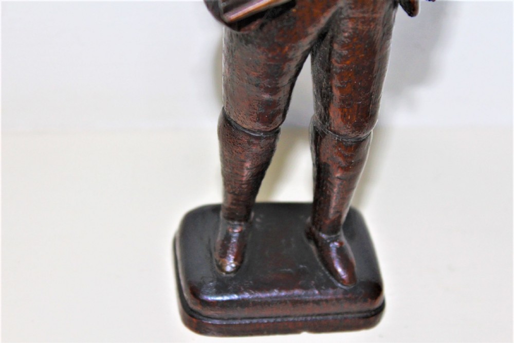 A hand-carved dark wood statuette depicting a male figure in 18th-century clothing holding a scroll on an integrated plinth. View V