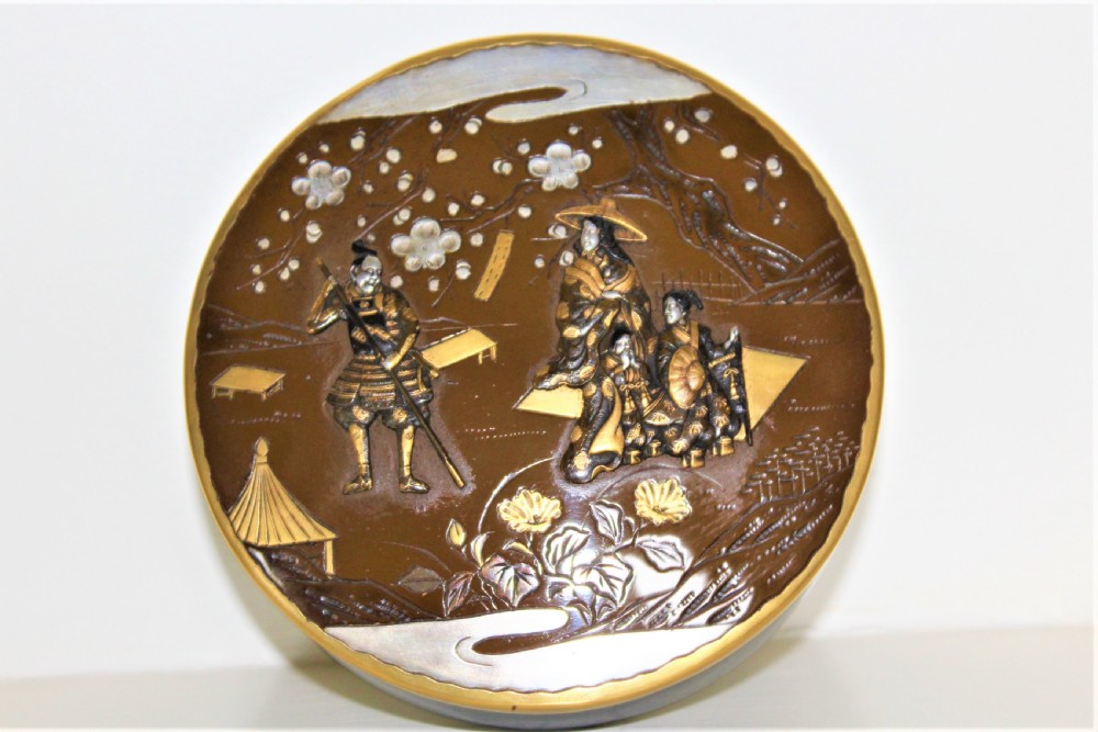 A circular brown bronze box lid decorated with high relief figures under cherry blossoms highlighted in gold and silver metal inlay.
