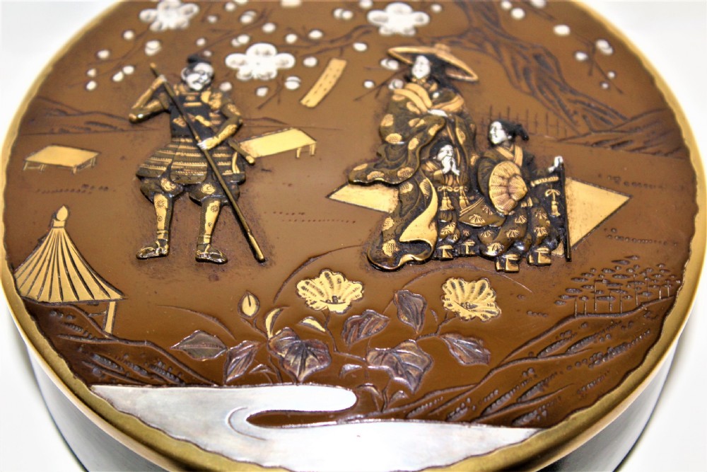 A circular brown bronze box lid decorated with high relief figures under cherry blossoms highlighted in gold and silver metal inlay. View III