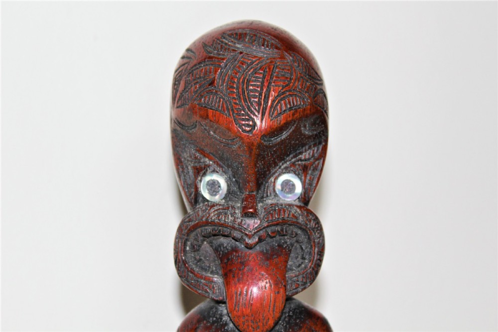 A carved reddish-brown hardwood standing figure featuring detailed facial tattoos, iridescent paua shell eyes, an extended tongue, and intricate carved body motifs. View III