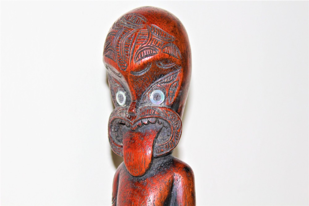 A carved reddish-brown hardwood standing figure featuring detailed facial tattoos, iridescent paua shell eyes, an extended tongue, and intricate carved body motifs. View IX