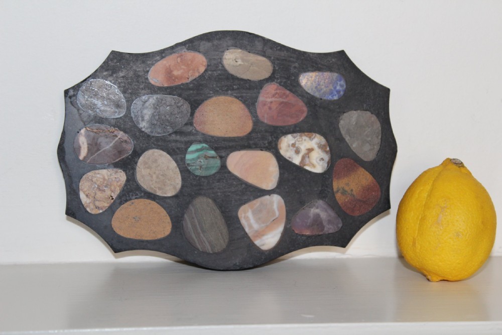 A shaped dark slate plaque inlaid with various polished colorful specimen stones including malachite and agates.
