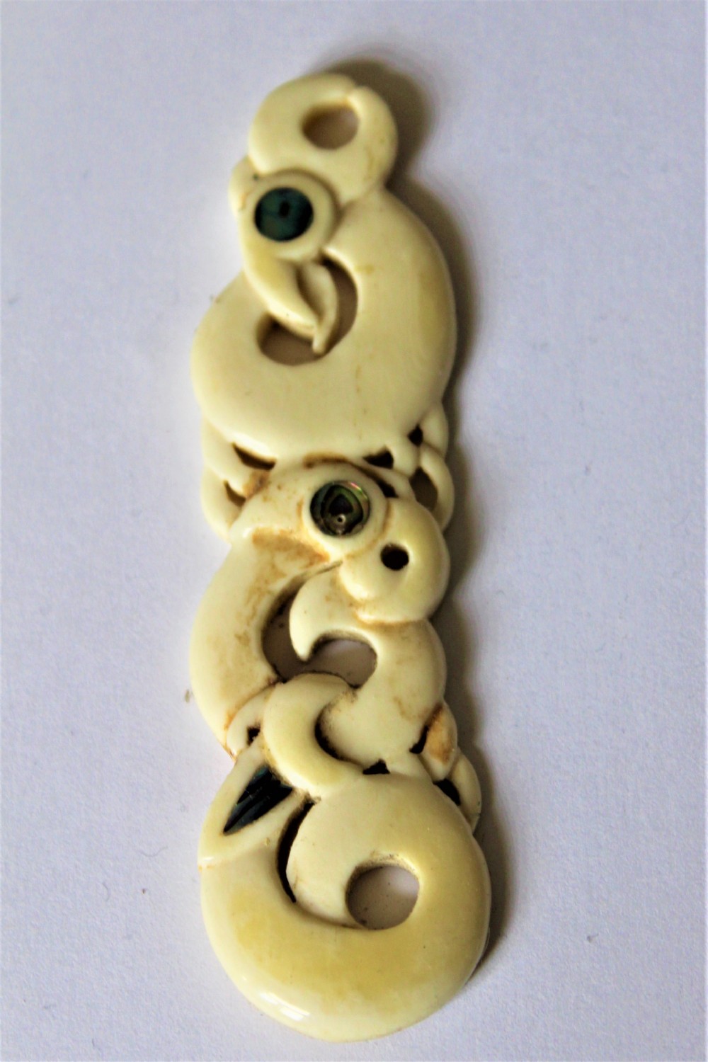 A carved cream bone pendant depicting interlocking stylized figures with inset green paua shell eyes.
