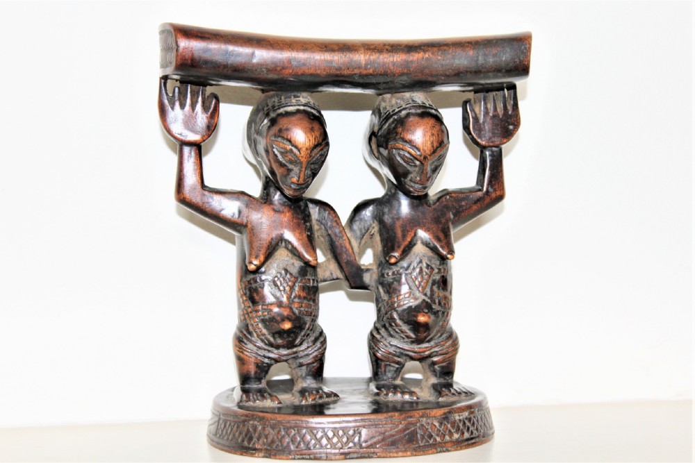 A carved dark hardwood headrest supported by two standing female caryatid figures on a circular carved base.