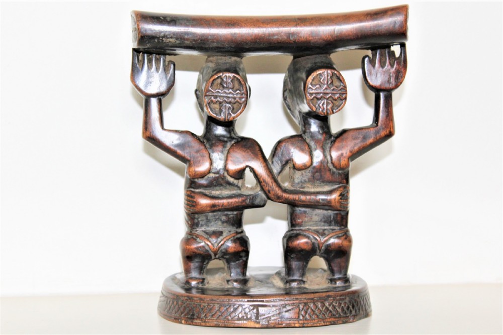 A carved dark hardwood headrest supported by two standing female caryatid figures on a circular carved base. View II