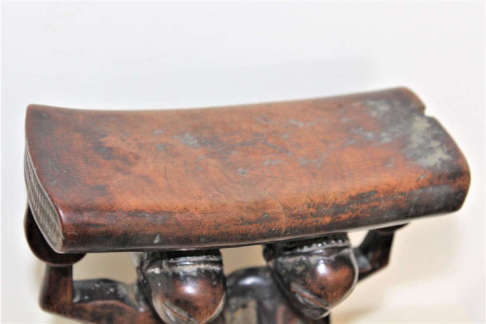 A carved dark hardwood headrest supported by two standing female caryatid figures on a circular carved base. View IX