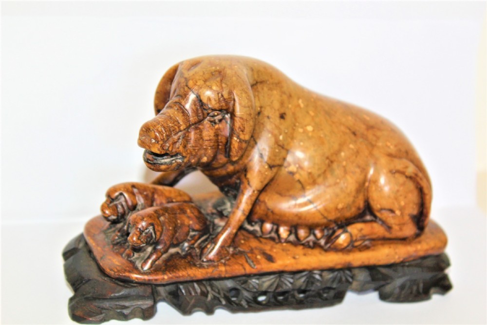 A carved warm brown soapstone sculpture depicting a sow reclining alongside her piglets on an ornate dark wooden stand.