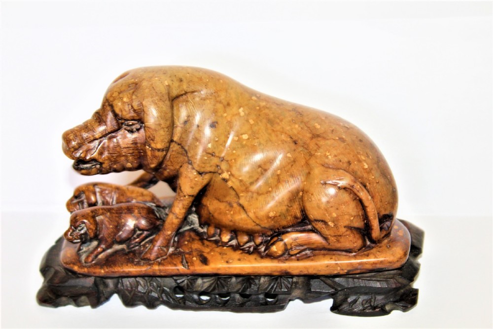 A carved warm brown soapstone sculpture depicting a sow reclining alongside her piglets on an ornate dark wooden stand. View II