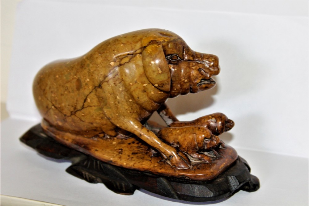 A carved warm brown soapstone sculpture depicting a sow reclining alongside her piglets on an ornate dark wooden stand. View VI
