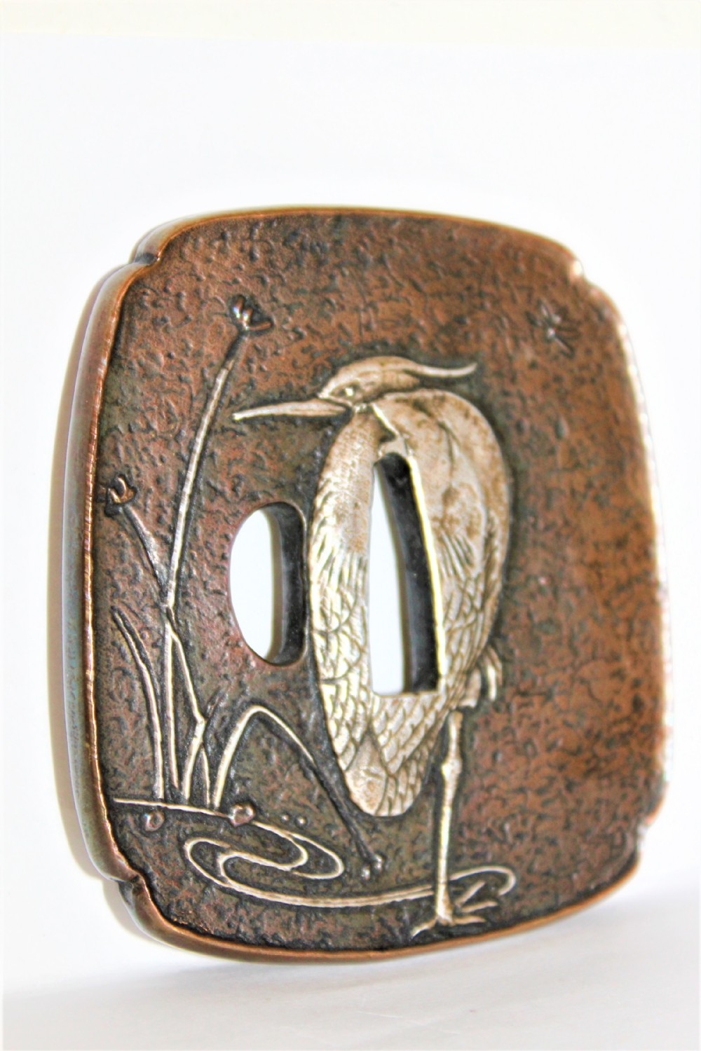 An antique dark bronze Japanese tsuba decorated with a silver overlay of a heron amongst water plants against a textured ground.