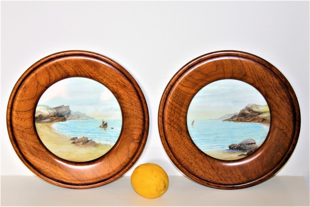 A pair of circular watercolour paintings depicting coastal landscapes with boats and cliffs, set inside polished wood circular frames.