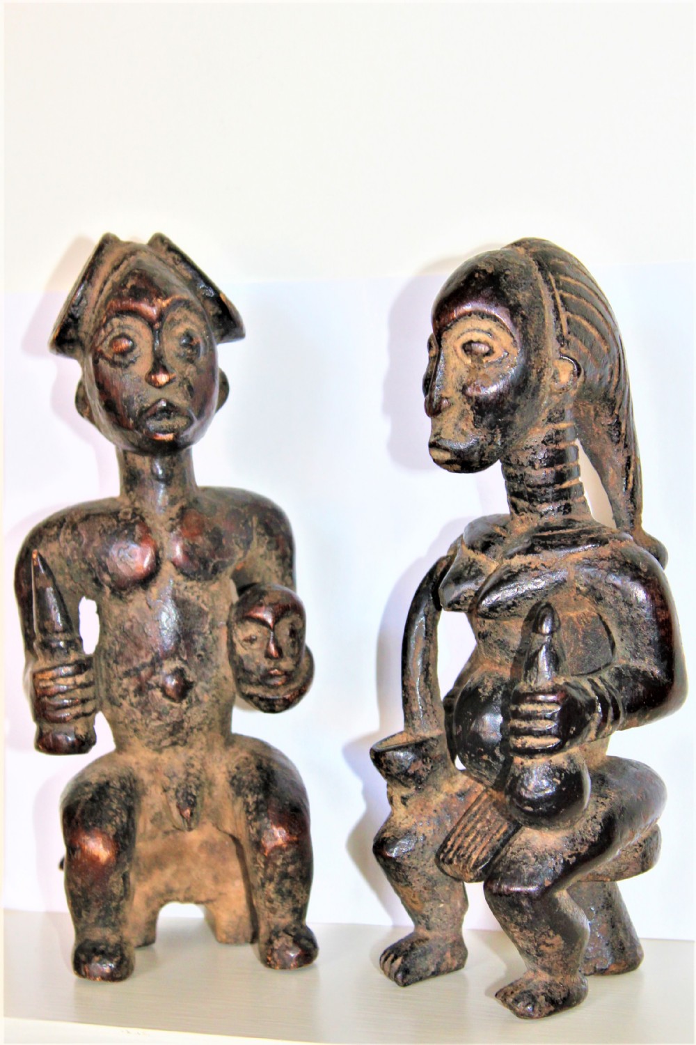 A pair of dark hand-carved wood Yoruba figurines depicting a male and female figure seated on low stools holding ritual items.
