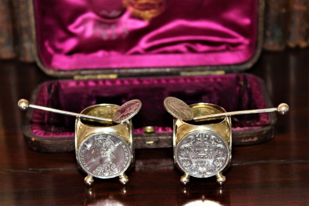 A pair of Victorian silver coin salt cellars with hinged coin lids and spoons sitting in front of an open fitted magenta velvet box.